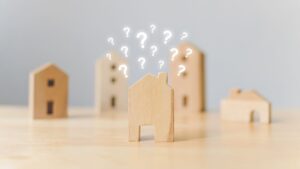 Wooden house models on a table, with glowing question marks above one house, symbolizing decision-making in real estate investment.