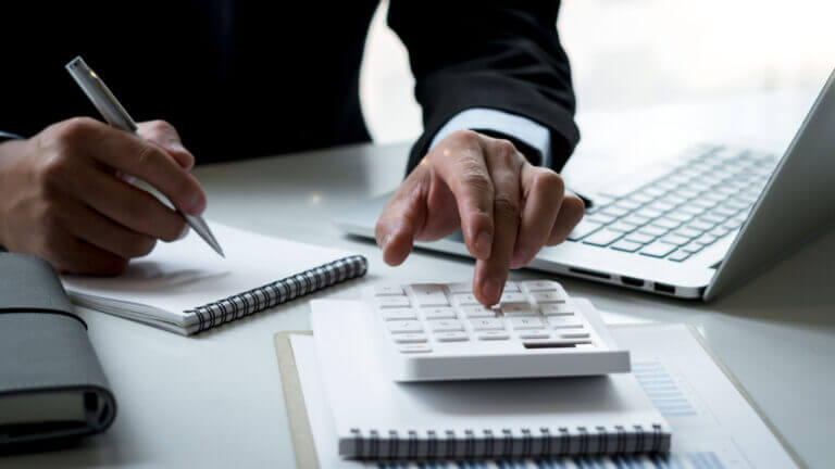 Close-up of a businessperson using a calculator at a desk, with a notepad, pen, and laptop, representing financial analysis and decision-making.